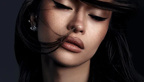 Woman with long black hair and closed eyes, wearing a chain necklace against a dark background.