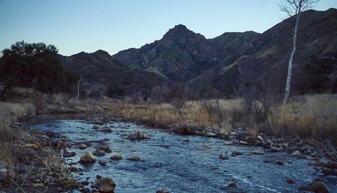 Mountain stream with rocks flowing through a valley with sparse winter vegetation and distant peaks.