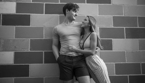 Couple posing against a brick wall, looking at each other with affection in black and white photography.