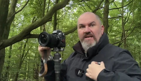 Photographer holding a professional DSLR camera on a tripod in a wooded forest setting.