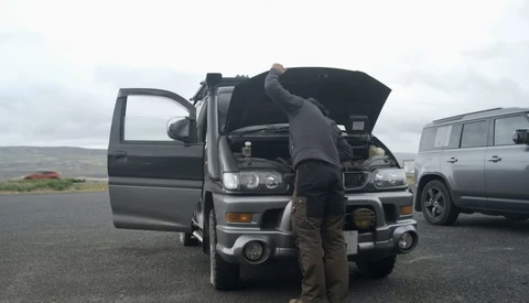 Photographer inspecting engine of parked vehicle during roadside shoot with overcast sky and mountains in background.