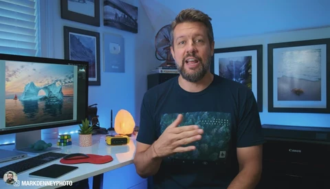 Man in dark t-shirt gesturing while speaking in a home office with blue ambient lighting and framed landscape photographs.