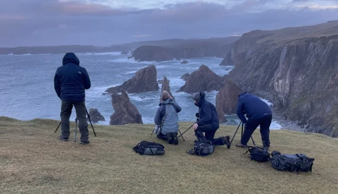 Photography crew of five setting up equipment on a coastal cliff overlooking dramatic rock formations and sea stacks.