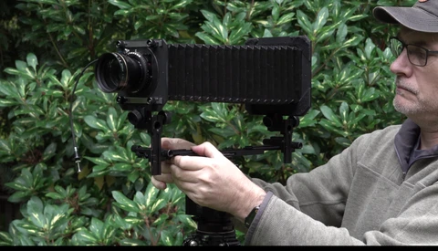Photographer holding a large format camera on a tripod while composing a shot against green foliage.