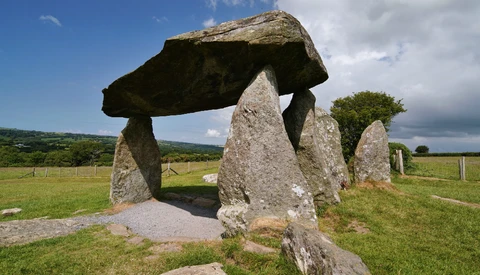 Ancient stone dolmen with large capstone balanced on upright stones in green pastoral landscape.