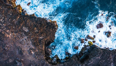 Aerial view of rocky coastline with waves crashing against dark stone cliffs.