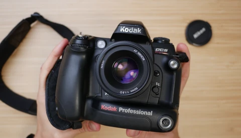 Hands holding a Kodak Professional DCS digital camera with attached grip and lens on wooden surface.