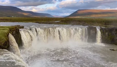 Powerful waterfall cascading over dark basalt cliffs into a turbulent river below, with mountains and moorland stretching across the background.