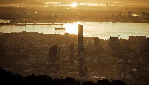 Golden-hour cityscape with dense urban skyline silhouetted against a bright waterway and hazy sky.