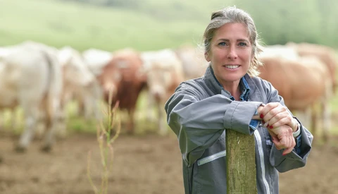 Woman in work clothes leaning on wooden fence with cattle grazing behind her.