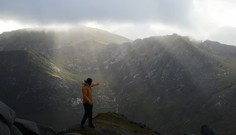 Hiker in yellow jacket standing on rocky ridge overlooking misty mountain valley.