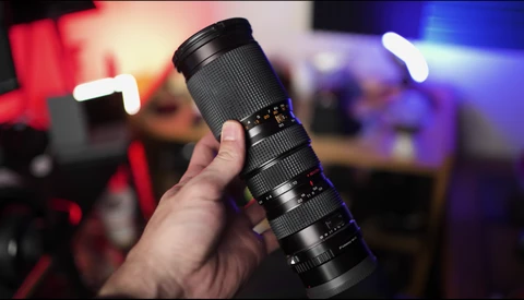 Hand holding a telephoto zoom lens with textured black barrel against blurred colorful background.