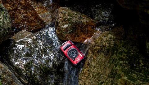 Red waterproof camera resting on moss-covered rocks beside flowing stream water.