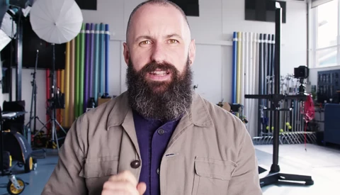 Bearded man in tan jacket gestures while speaking in a photography studio.