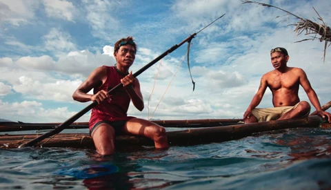 Two shirtless men in shallow water with wooden poles and a traditional boat.