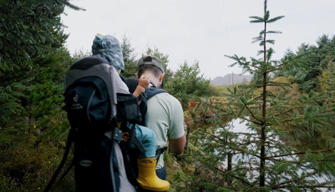 Backpacker wearing large hiking pack walks along forested trail beside calm water.