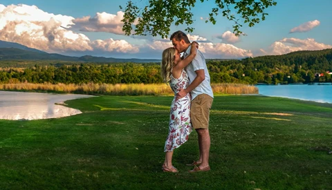 Couple kissing in a grassy field with mountains and lake in the background.