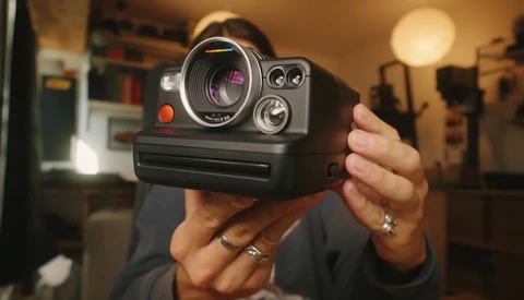 Person holding a vintage instant camera with manual controls and a purple-tinted lens element visible.