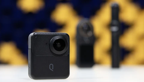 Compact black action camera with circular lens mounted on white surface, with blurred camera equipment in background.