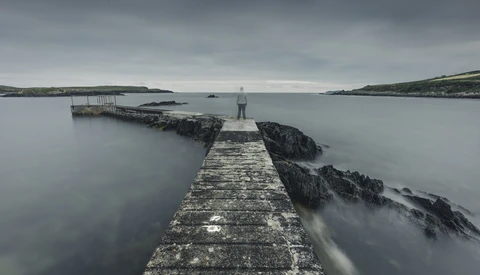 Long stone pier extending into calm gray water with rocky outcrops and distant coastline under overcast sky.