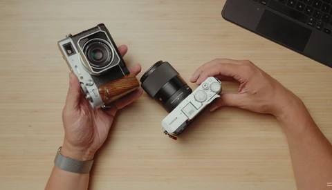 Hands holding two vintage rangefinder cameras with interchangeable lenses on a wooden desk.