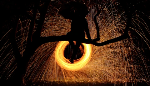 Silhouetted figure spinning steel wool in a circular motion, creating a bright golden ring of sparks against black surroundings.