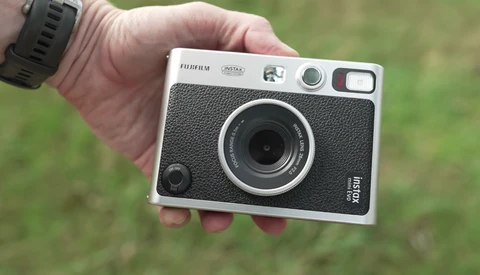 Hand holding a Fujifilm Instax hybrid instant camera with a black textured body and silver trim against a blurred green background.