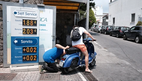 Person bending over a blue Vespa scooter parked in front of a gas station price sign on an Italian street.