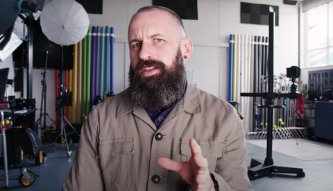 Bearded man in tan work shirt gesturing while speaking in a professional studio setting.