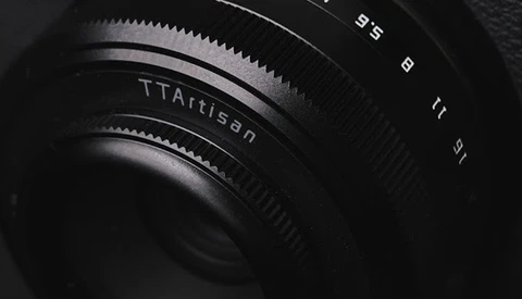 Close-up of a TTArtisan camera lens showing focus ring and aperture markings in dramatic side lighting.
