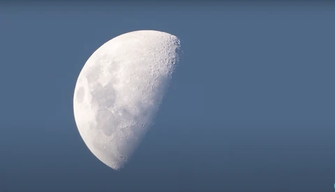 Waning crescent moon against a clear blue sky, surface details and craters visible in sharp focus.