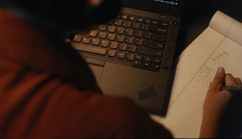 Person in rust-colored sleeve pointing at handwritten notes beside a laptop keyboard during evening work session.