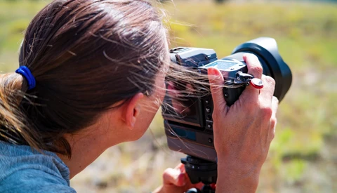 Photographer holding a video camera to their eye while operating it outdoors in daylight.