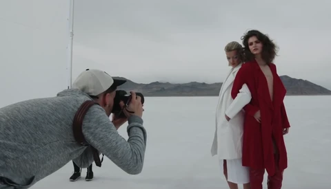 Photographer in white cap and gray jacket shooting two women in red and white clothing standing by a calm body of water.