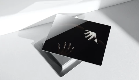 Black card with white hands reaching toward each other, positioned on white geometric surfaces with dramatic diagonal lighting.