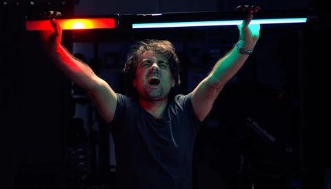 Man holding illuminated neon tubes overhead with arms raised in celebration against dark background.