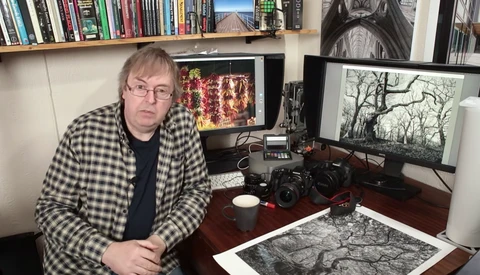 Photographer at desk surrounded by cameras, monitors displaying landscape images, and film prints.
