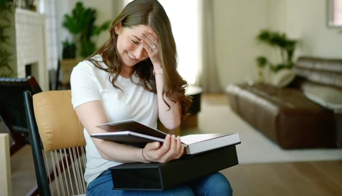 Woman sitting indoors holding stacked books with hand to forehead, appearing stressed or overwhelmed.