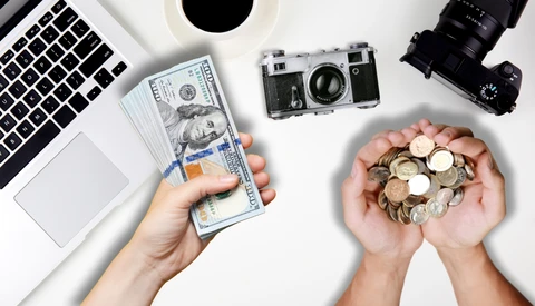 Hands holding US currency and coins next to vintage cameras and laptop on white surface.