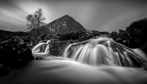 Long-exposure photograph of a rushing mountain stream with a bare tree and rocky peak in the background.