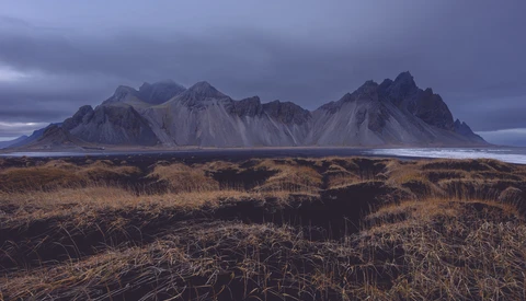 Dramatic mountain range with dark peaks rising above golden tundra landscape under moody twilight sky.