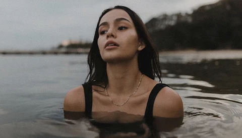 Woman in black swimsuit partially submerged in calm water, gazing upward with a contemplative expression.