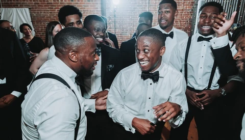 Men in formal attire laughing and conversing at an indoor event with exposed brick walls.