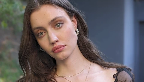 Woman with long dark hair tilting her head slightly, wearing pearl earrings and a delicate chain necklace.