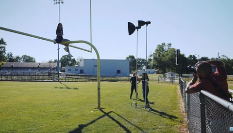 Two people standing on a sunny baseball field with tall light poles and a blue building visible in the background.