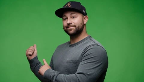 Man wearing black cap and gray long-sleeve shirt giving a thumbs-up gesture against green background.