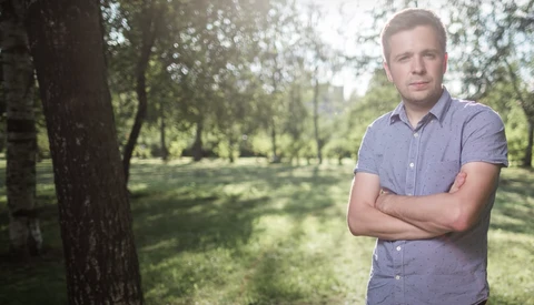Man in light purple shirt standing with crossed arms in a park with tall trees and dappled sunlight.