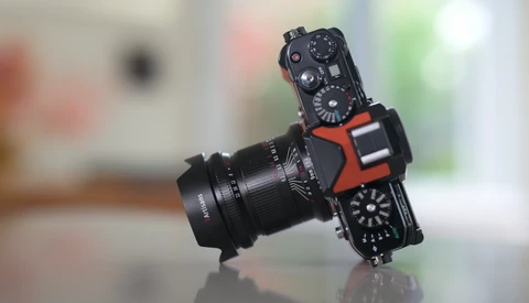 Black mirrorless camera with red accent grips and vintage manual focus lens mounted, photographed against blurred indoor background.