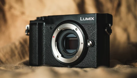 Panasonic Lumix mirrorless camera body with lens mount exposed, positioned on sandy surface with warm-toned background.