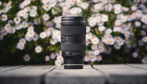 Black telephoto zoom lens standing upright on concrete with soft-focus daisies in background.